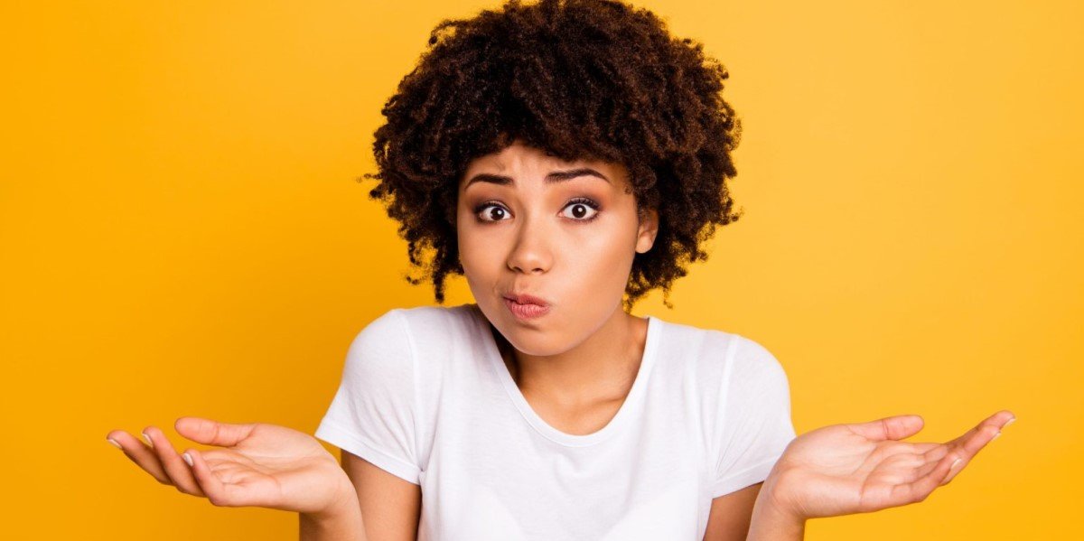 Jonge vrouw met donker gekruld haar trekt haar schouders op, handen open, mimiek oeps foutje, voor geel achtergrond